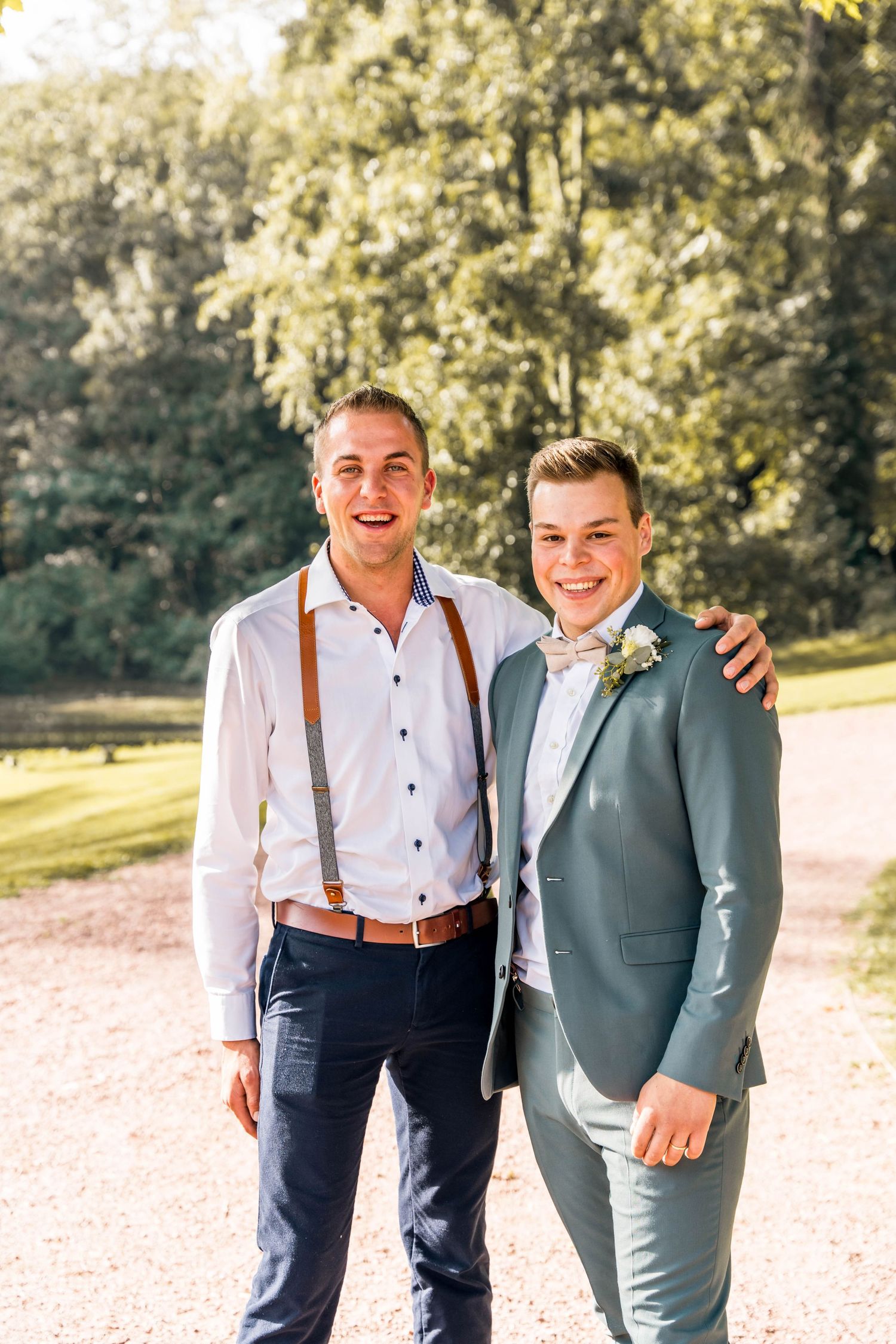 Two groomsmen in formal attire smiling on sunlit garden path.