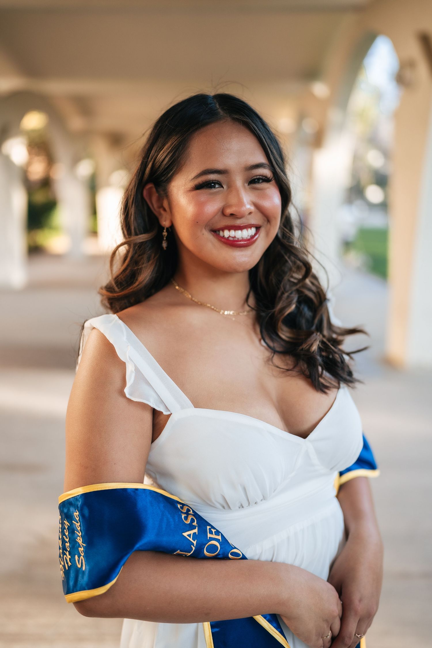 A graduate wearing a white dress and blue sash poses under an outdoor colonnade with a bright smile.