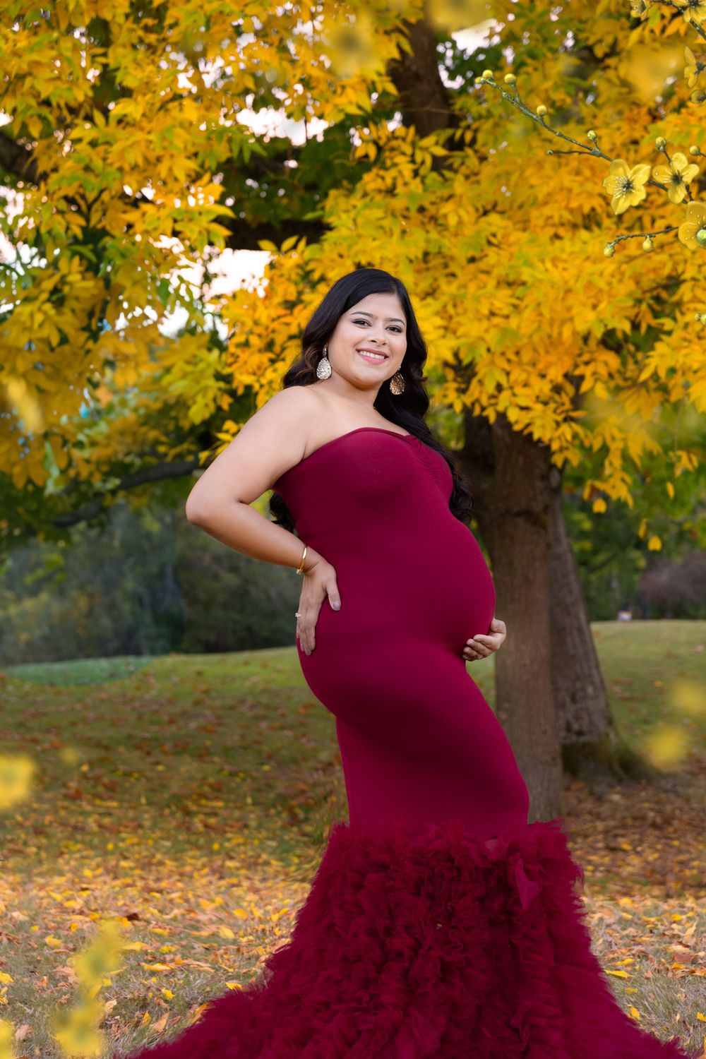 Maternity photoshoot outdoors with burgundy mermaid-style dress against vibrant yellow autumn foliage.