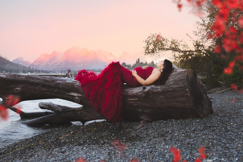 Dreamy scene of someone in a flowing red dress reclining on driftwood by a misty beach at sunset.