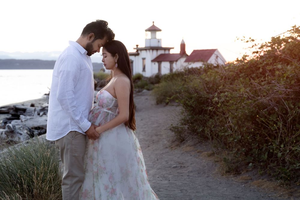 A couple embraces on a coastal path near a lighthouse at sunset, with water and rocky shoreline in the background.