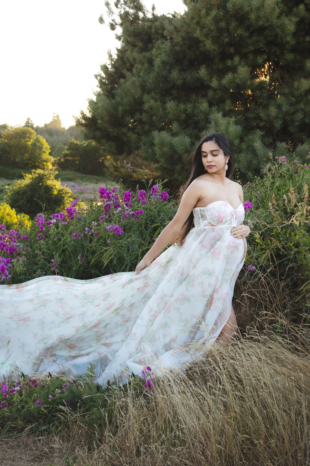 A flowing white floral gown cascades over dry grass near purple wildflowers and evergreen trees at sunset.