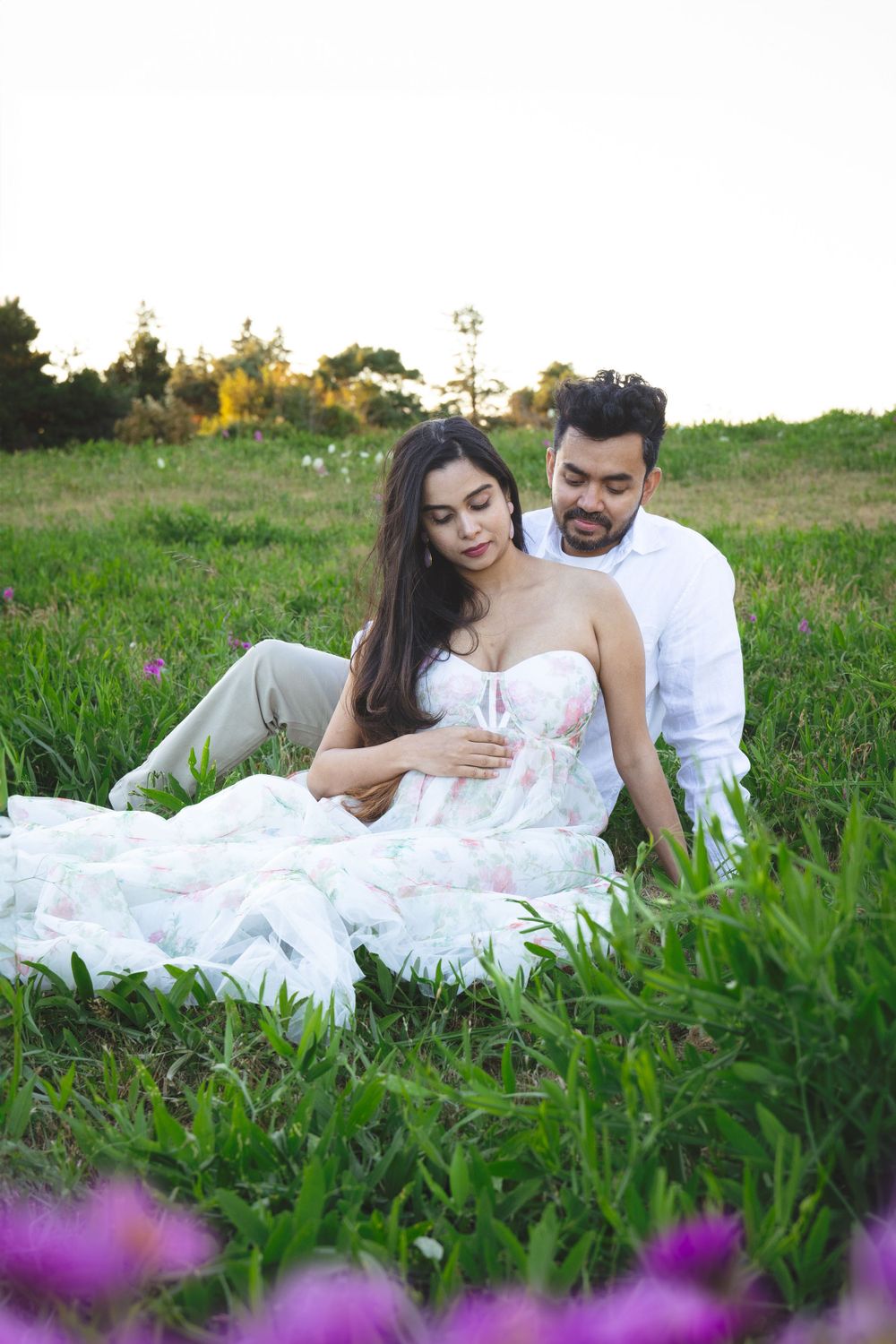 A couple in romantic white attire sits together in a meadow with purple wildflowers in the foreground.
