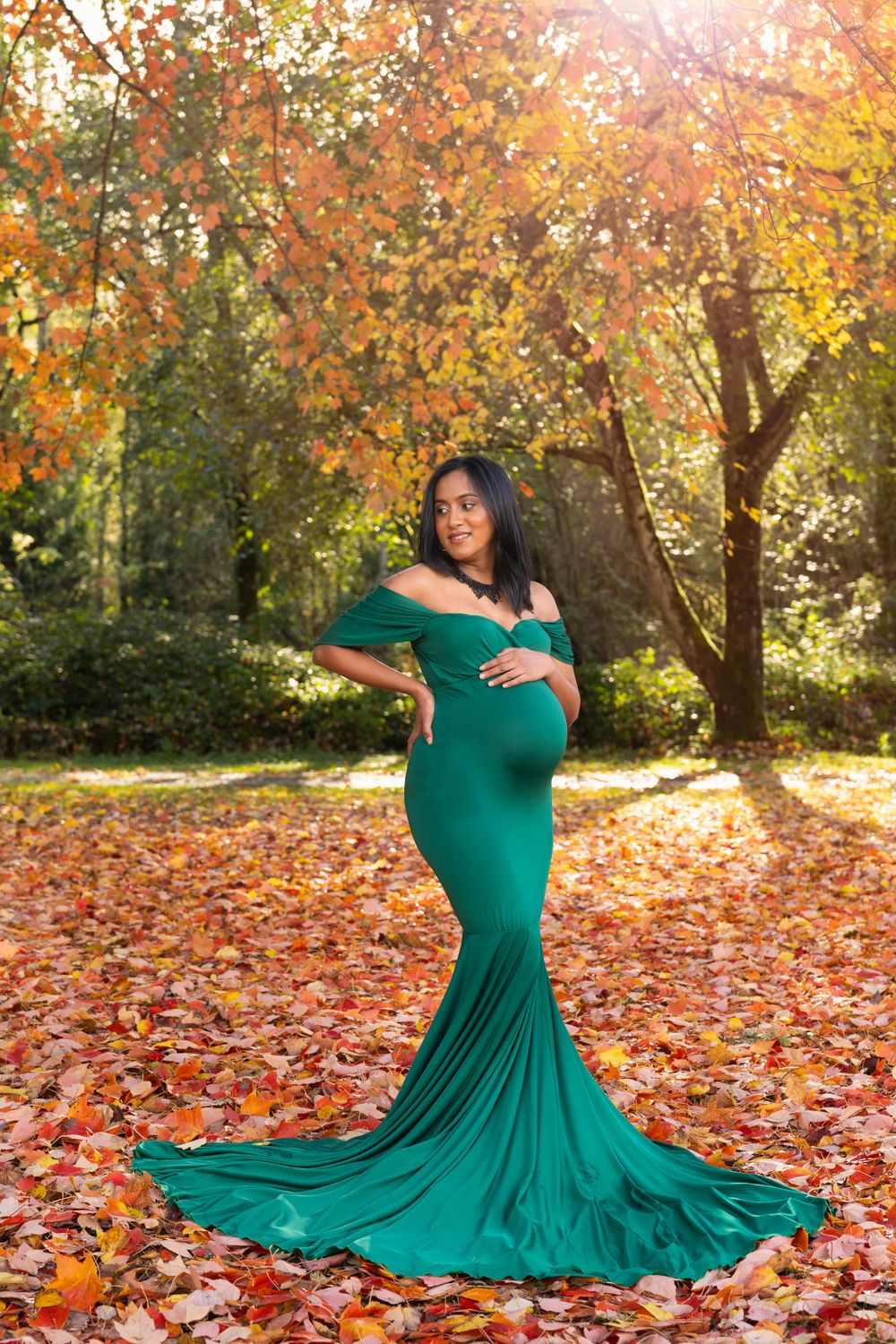 An elegant emerald green maternity gown with flowing train rests on colorful autumn leaves beneath golden trees.