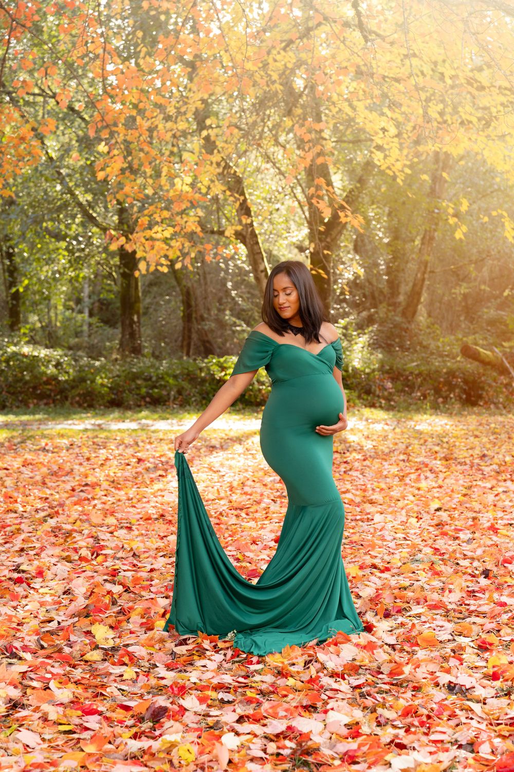 Elegant maternity photo in emerald green dress surrounded by vibrant autumn leaves and golden sunlight in forest setting.
