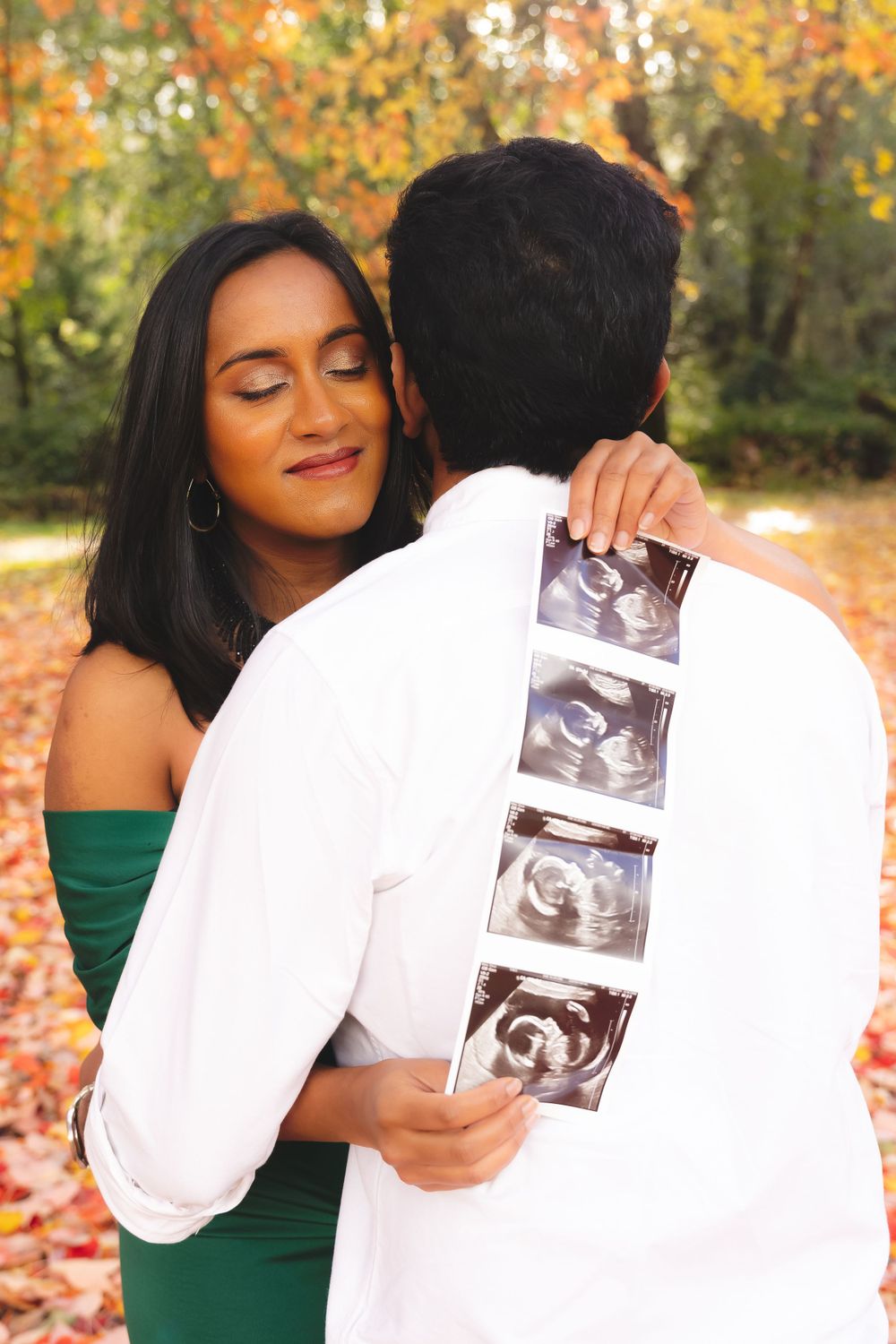 A couple embraces outdoors in autumn foliage while holding ultrasound photos, celebrating their pregnancy announcement.