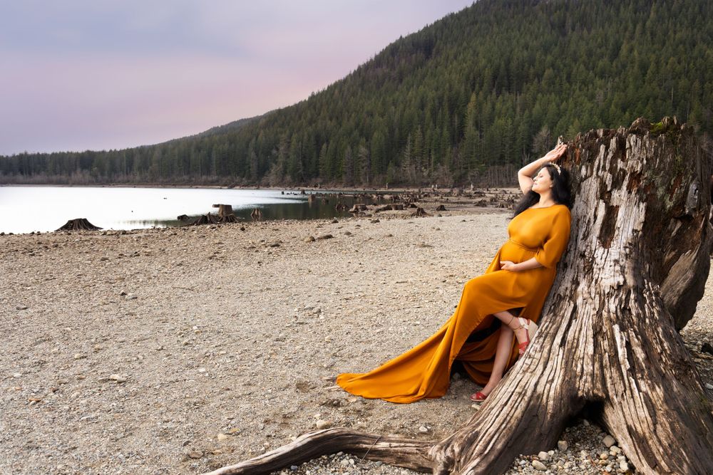 A person in a flowing yellow dress reclines against a driftwood log on a remote beach with forested mountains in background.