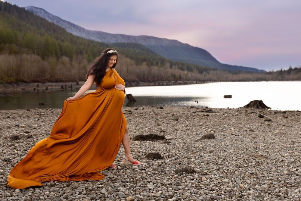 A flowing orange dress billows in the wind against a scenic mountain lake backdrop at dusk.