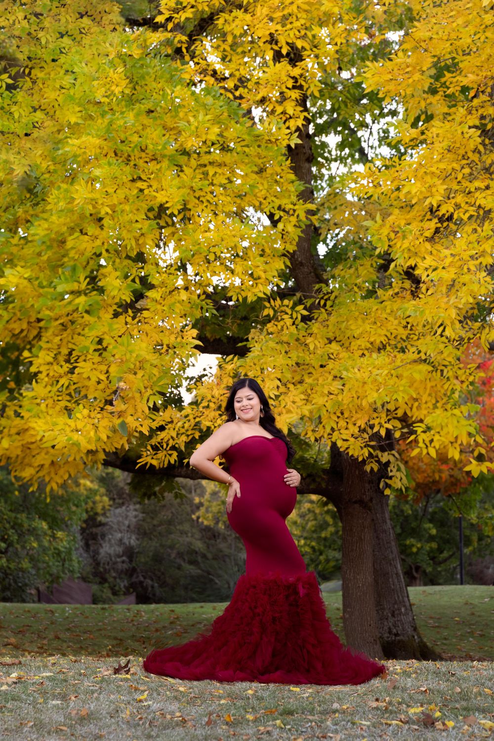 A burgundy maternity gown dress contrasts beautifully against vibrant yellow fall foliage in an outdoor portrait setting.