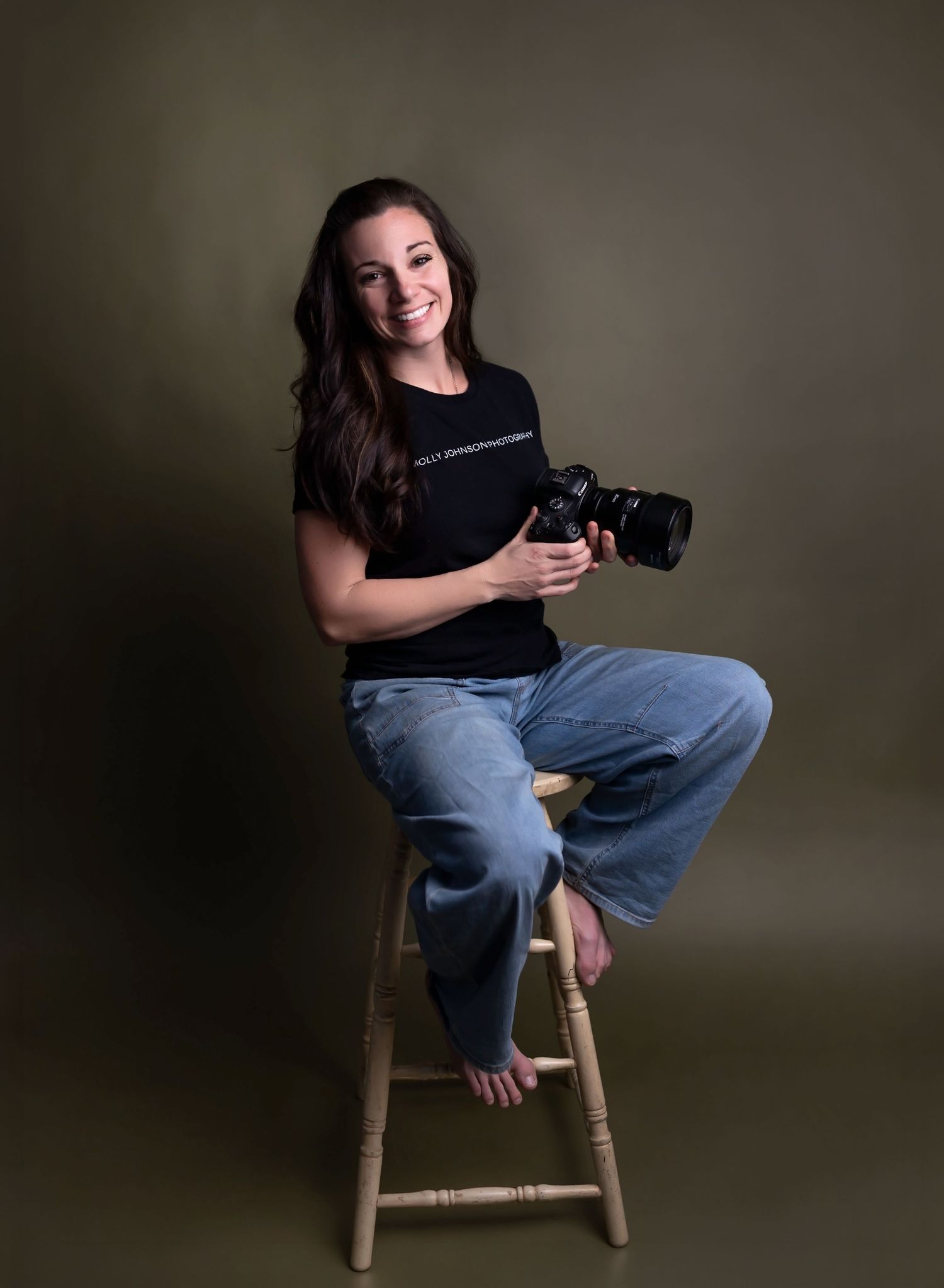 A photographer in a black shirt and jeans sits casually on a wooden stool while holding a professional camera.