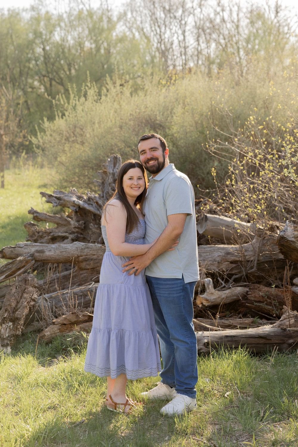 A couple stands together embracing outdoors in front of fallen logs during golden hour lighting at river bends park in utica michigan