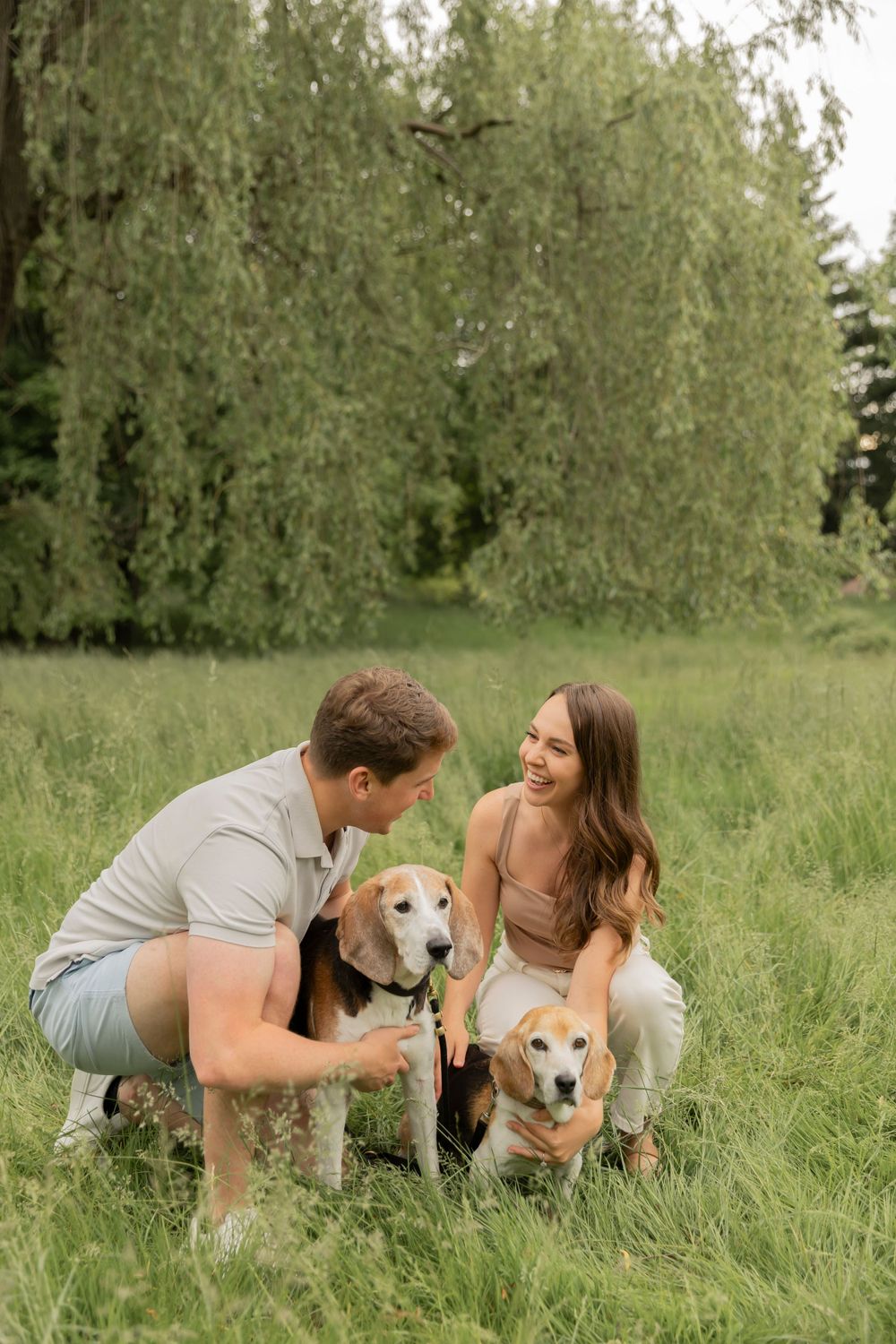couple crouching down by their dogs in a grasses field and smiling at each other