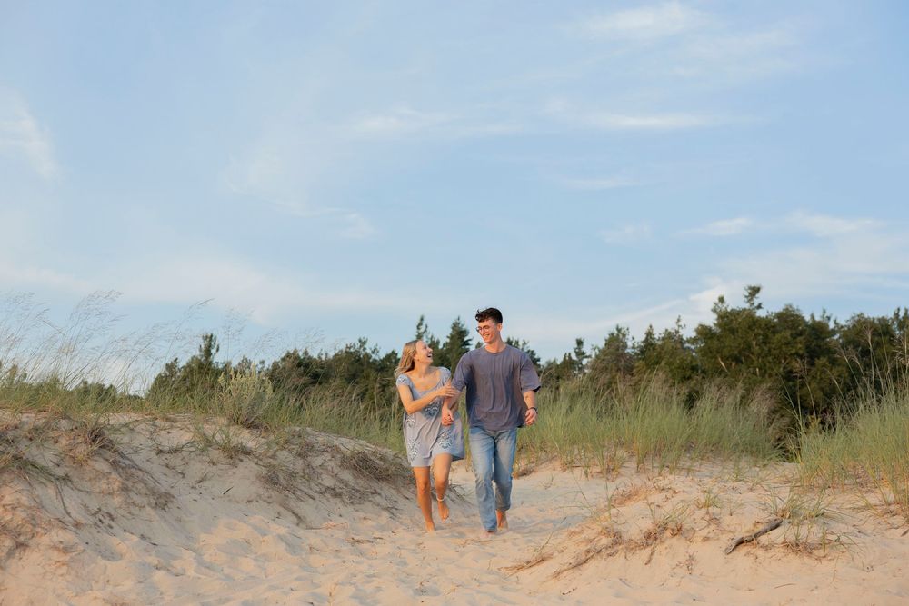 Couple walking hand in hand on sandy beach dunes with green trees and blue sky in background.
