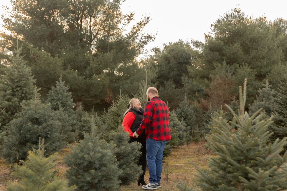 couple wearing buffalo plaid smiling at each other in a field of evergreen trees in lake Orion michigan
