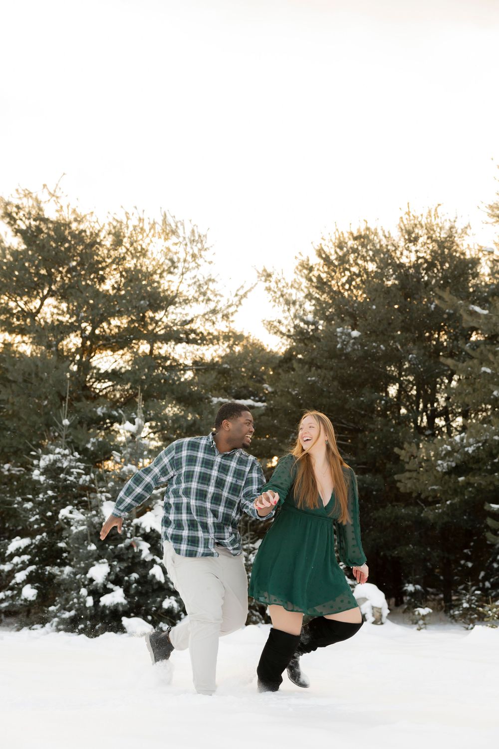 A couple in winter attire shares romantic moments in a snowy landscape with evergreen trees at sunset at Addison oaks Christmas tree farm