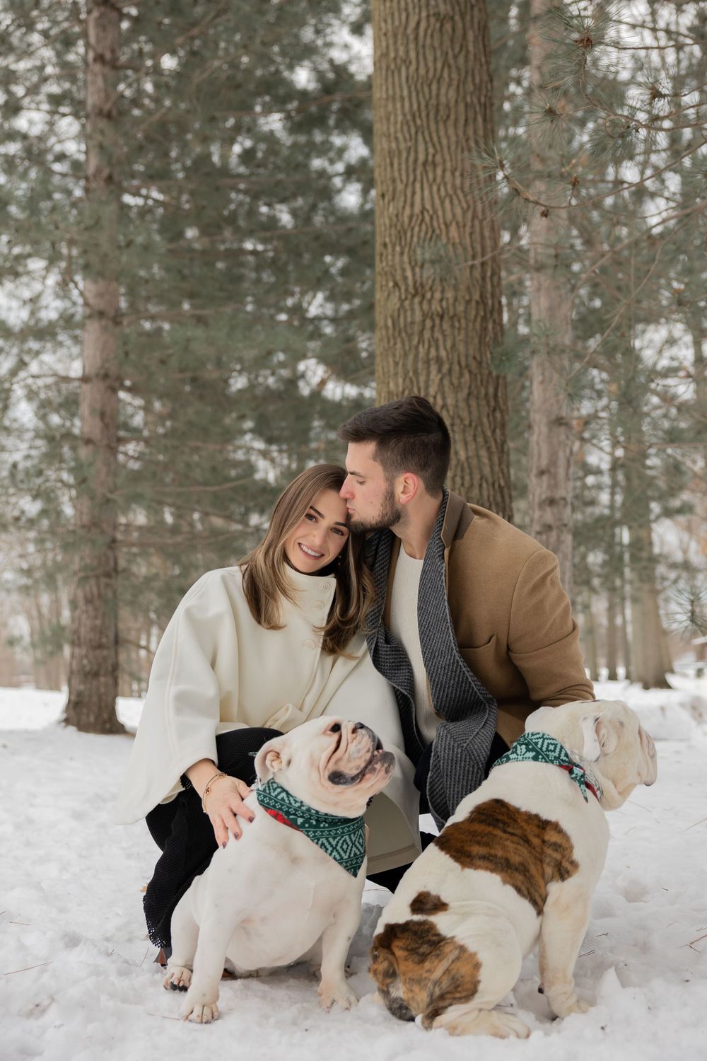 A couple sits in snowy winter forest with their two dogs enjoying an intimate outdoor photo session at firefighters park in troy michigan