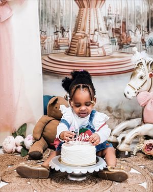 A toddler girl sits on a rug before a white birthday cake, surrounded by stuffed animals and a carousel-themed backdrop.