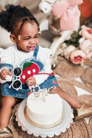 Toddler girl smiling at her first birthday cake with One topper, wearing denim strawberry dress.