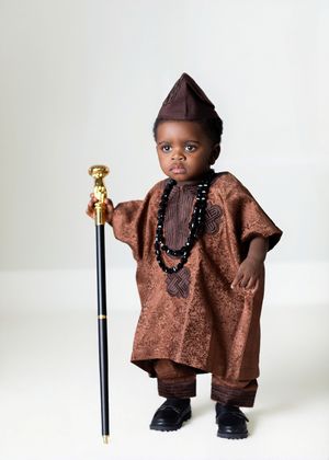 Toddler in traditional African brown agbada outfit with matching fila hat, beaded necklace, and gold-topped cane.