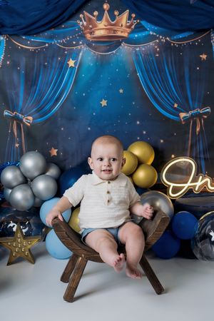 First birthday photo setup with blue draped backdrop, crown decor, balloons and wooden chair against starry theme decorations.