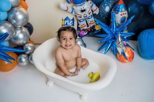 Small child sits in white bathtub with yellow rubber duck surrounded by blue and silver space-themed party decorations.