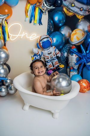 Festive first birthday cake smash photo session with blue and silver balloons surrounding baby in white bathtub.