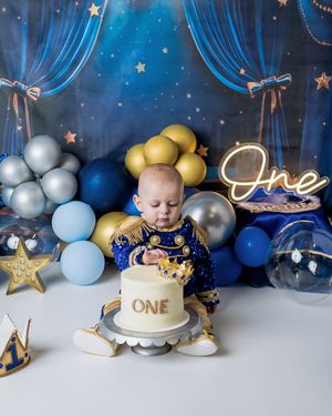 First birthday setup with blue and gold balloons, stars, blue drapes, and a small cake on a white pedestal against dark backdrop.