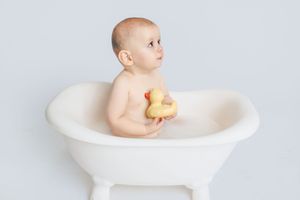 Baby plays with rubber duck while sitting in white bathtub during bath time.