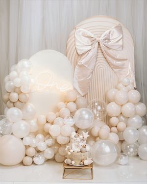Elegant first birthday balloon garland display with cream and white balloons, neon 'One' sign, and large satin bow backdrop.