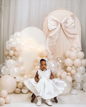 Baby girl in white dress and bonnet sits at her first birthday party with balloon arch and neon One sign.