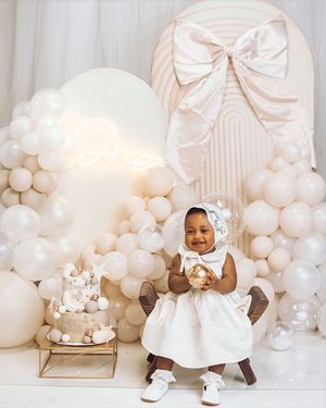A smiling baby girl in a white dress celebrates her first birthday surrounded by white balloons and a decorated cake.