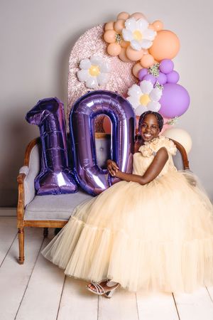 Birthday celebration setup with purple number 10 balloons, floral decorations, and soft cream tulle dress against gray backdrop.