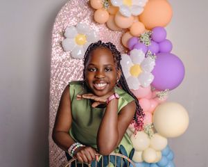 Young child poses in front of a pink backdrop with colorful balloon garland featuring peach, purple and ivory balloons.