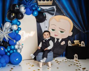 A baby boy in a suit sits at a Boss Baby themed first birthday party with blue and black balloons.