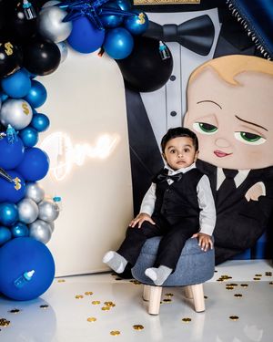 Baby boy in black suit sits at Boss Baby themed first birthday party with blue balloons and gold confetti.