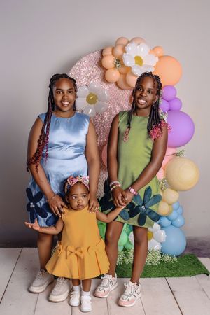 Three people stand in front of a pastel balloon arch and floral backdrop wearing blue, yellow and green dresses.