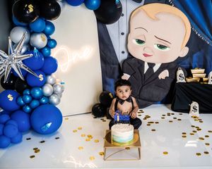 Baby's first birthday smash cake with Boss Baby theme, blue balloons, gold confetti, and decorative backdrop.