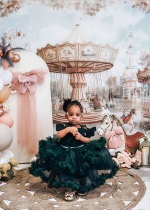 Toddler girl in dark green tulle dress poses before a carousel backdrop with balloons and floral decorations.