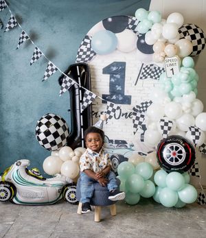 Toddler boy smiles at race car themed first birthday party with checkered flags, balloons, and number one decorations.