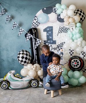 Two boys pose at a race car themed first birthday party with checkered flags, balloons, and a number one backdrop.
