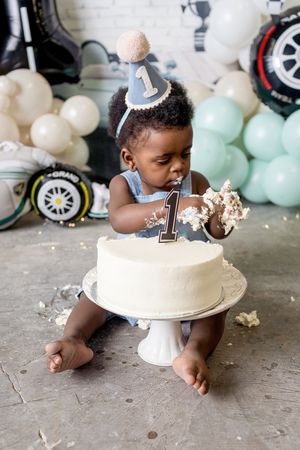 Baby's first birthday cake smash with a number 1 hat, white frosted cake, and balloon decorations in blue and white.
