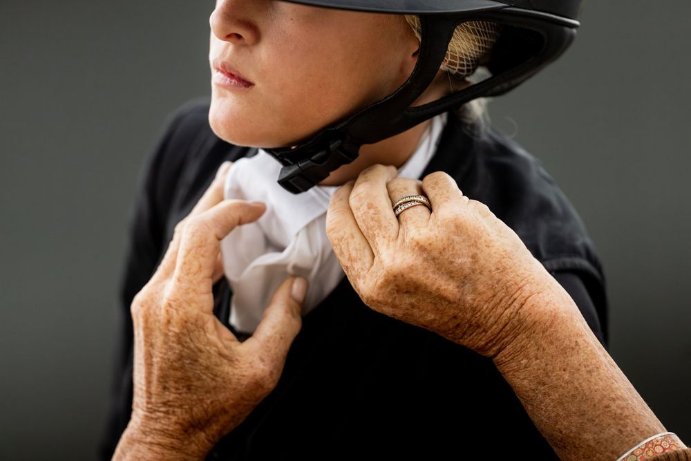 Elderly hands adjusting a black equestrian helmet chin strap on a young woman wearing a white stock tie.