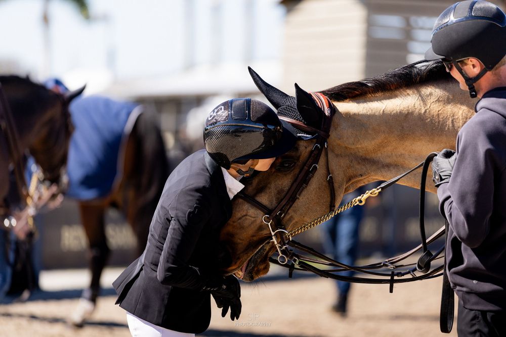 Equestrian rider in black helmet and jacket affectionately nuzzling a brown horse at an outdoor competition.