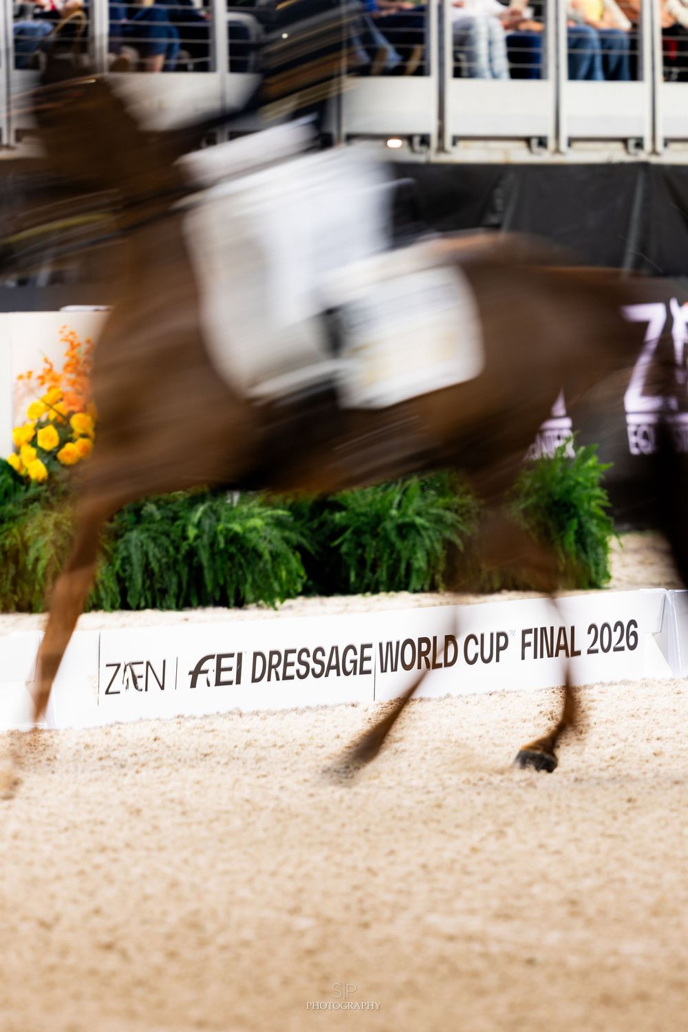 A horse and rider blur past the FEI Dressage World Cup Final 2026 arena barrier in a dynamic motion-blur action shot.