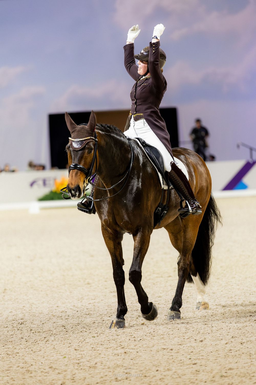 Female equestrian rider on bay horse raises hands in victory during dressage competition in sandy arena.