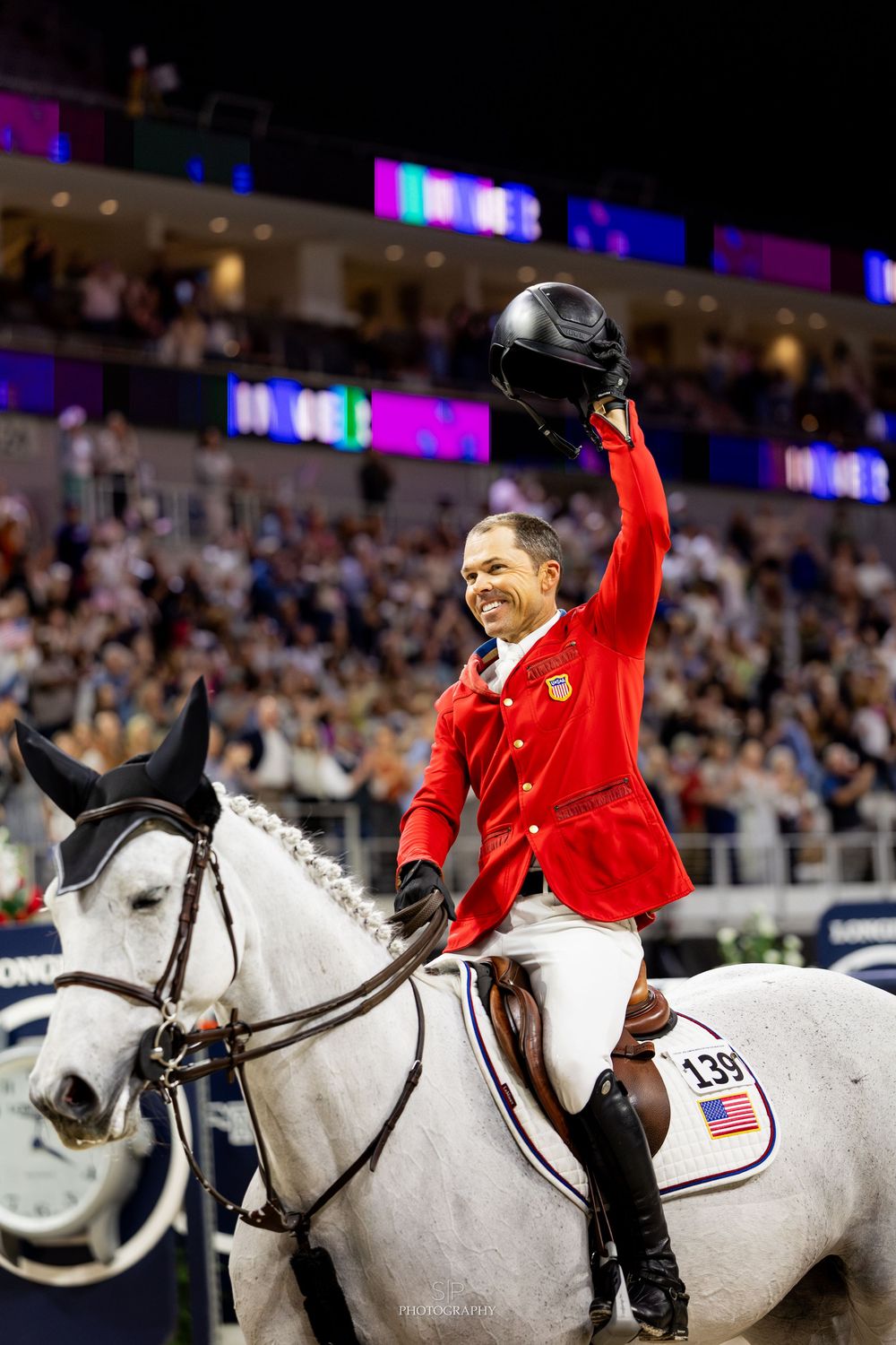 Equestrian rider in red jacket celebrates victory on white horse, raising helmet to cheering crowd at indoor arena.