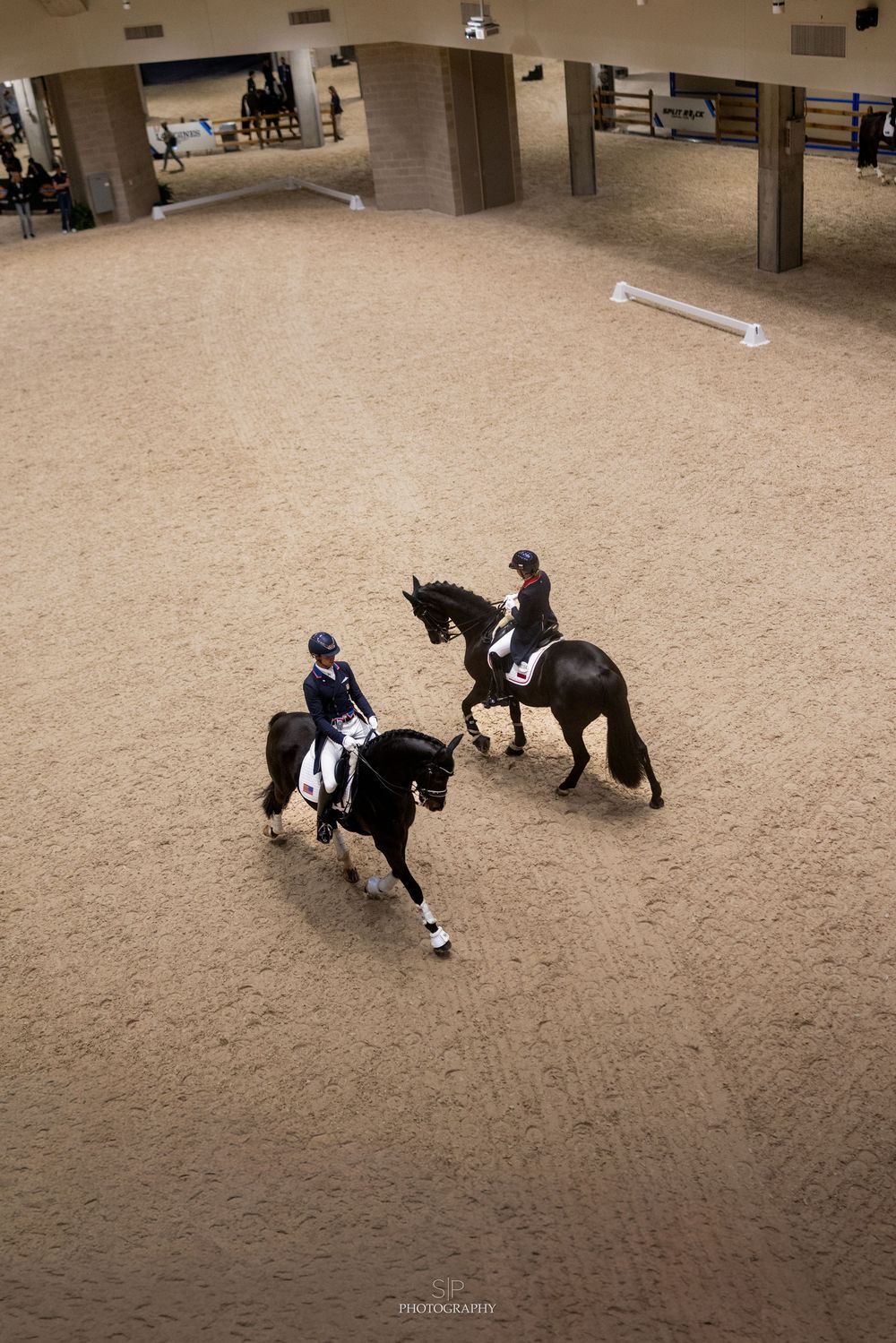 Two riders on black horses practice dressage in an indoor sand arena, viewed from above.
