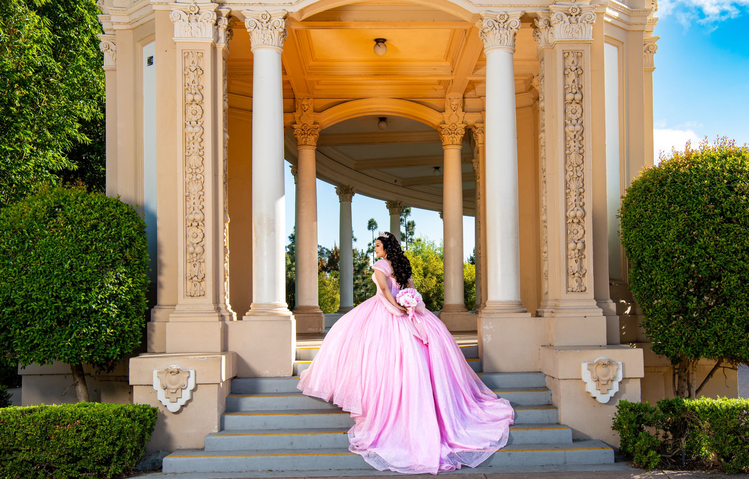 A flowing pink ballgown cascades down marble steps at an elegant columned pavilion with manicured hedges under blue skies.