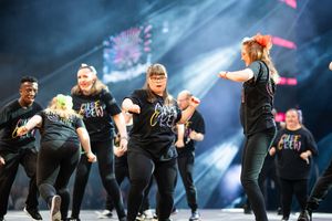 Performers in black outfits dance in synchronized formation on stage with dramatic blue lighting effects.
