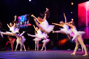 Ballet dancers in white tutus perform graceful movements on a colorfully lit stage during a dance recital.